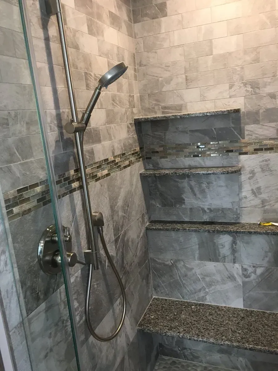 Gray-tiled shower with built-in shelves, handheld showerhead, and glass door.