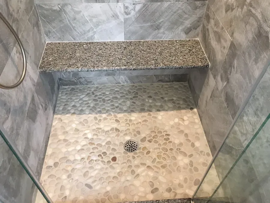 Shower with pebble tile floor, granite bench, gray tile walls, and glass enclosure.
