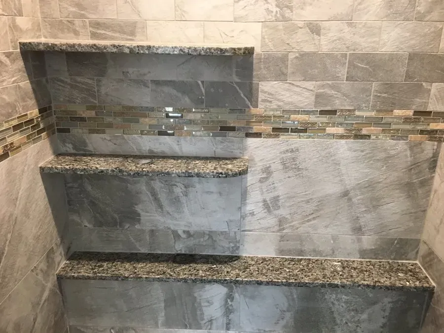 Bathroom shower wall with three built-in shelves, gray and tan tile with granite edges, and a decorative mosaic strip.