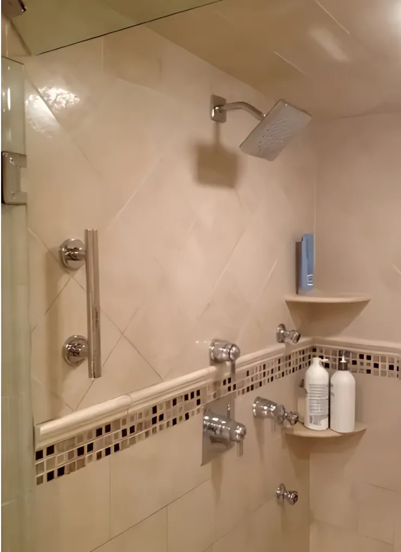 Shower with beige tile, mosaic trim, and chrome fixtures; a rectangular showerhead and a shelf with toiletries.