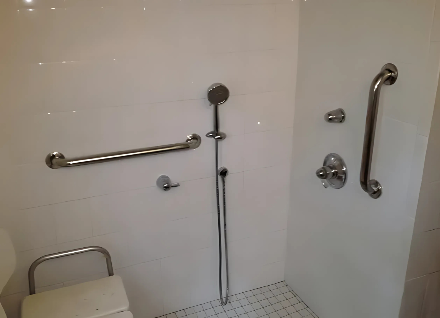 Accessible white tiled shower with grab bars, a shower seat, and handheld shower head.