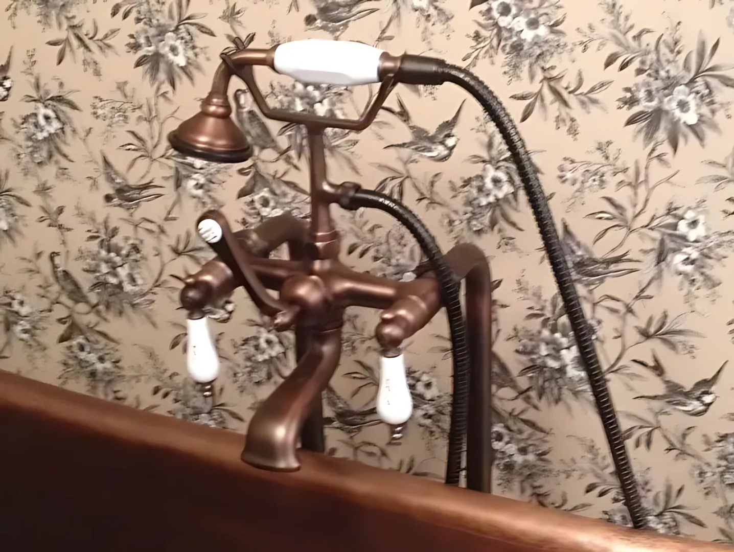 Copper bathtub faucet with white handles and a handheld shower against floral wallpaper.