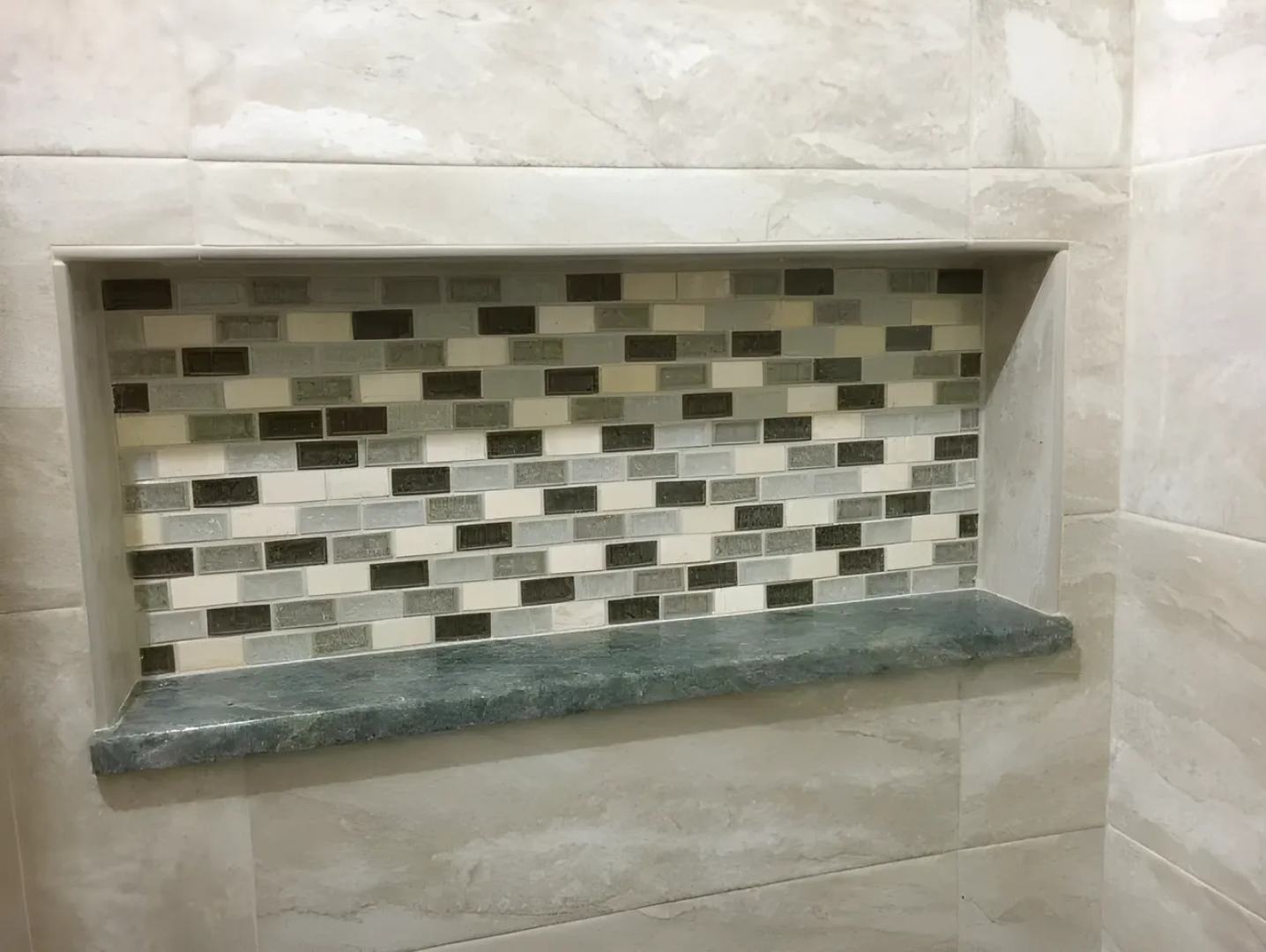 Shower niche with a stone shelf, gray, white, and dark gray mosaic tile backing.