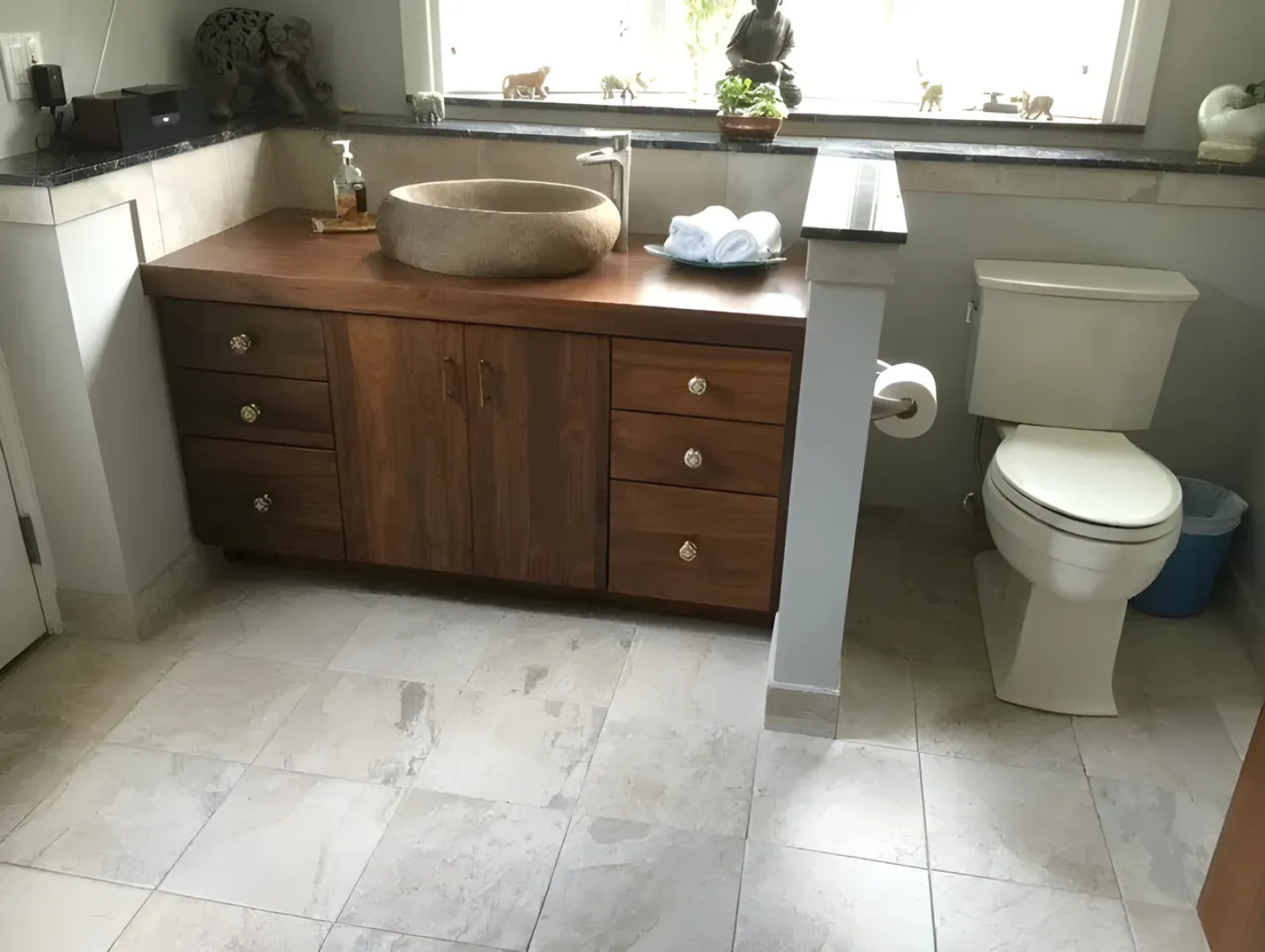 Bathroom with a wooden vanity, stone sink, toilet, and light-colored tiled floor.