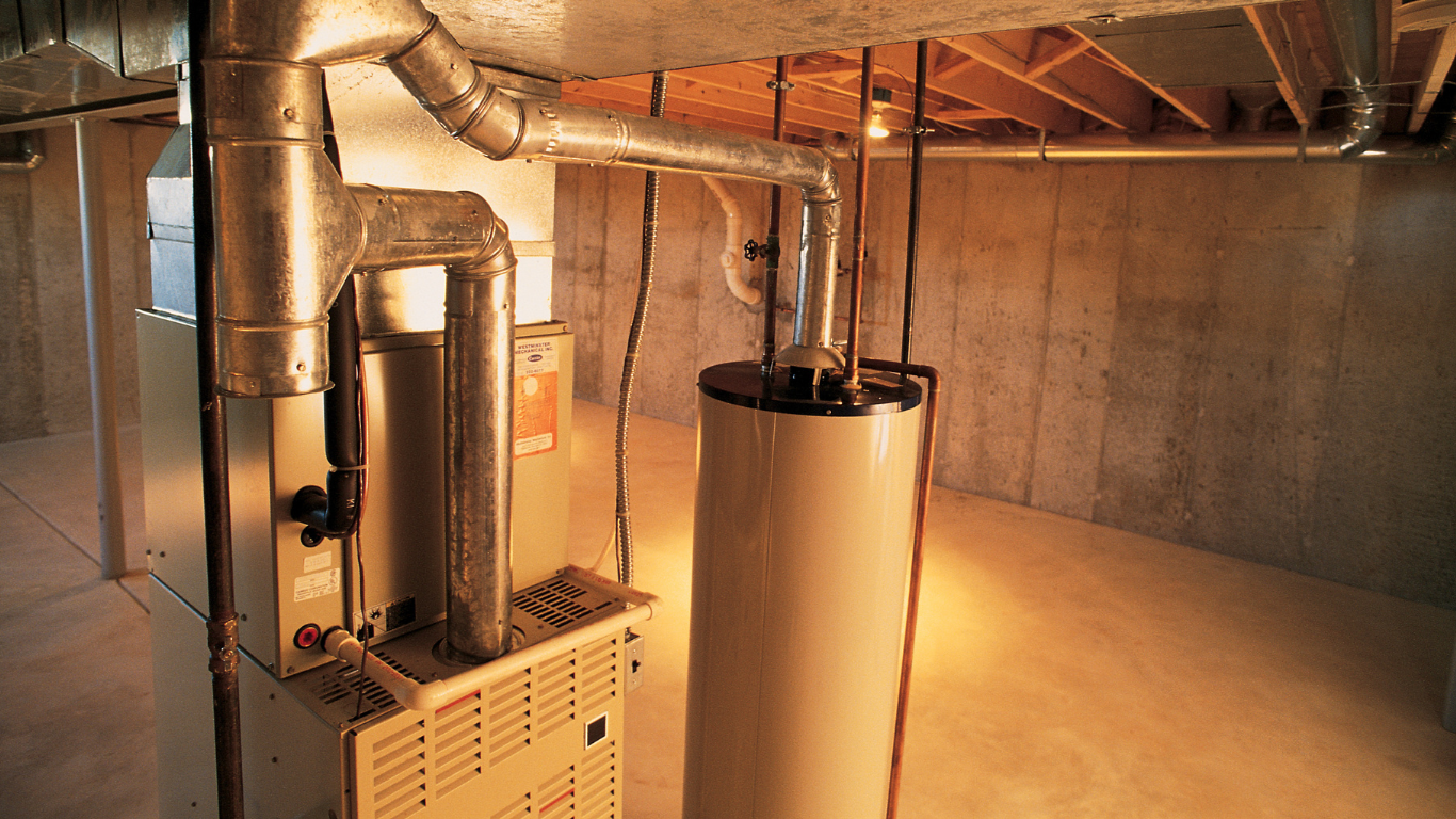 Furnace and water heater in a basement. Tan metal units with ductwork against concrete walls.