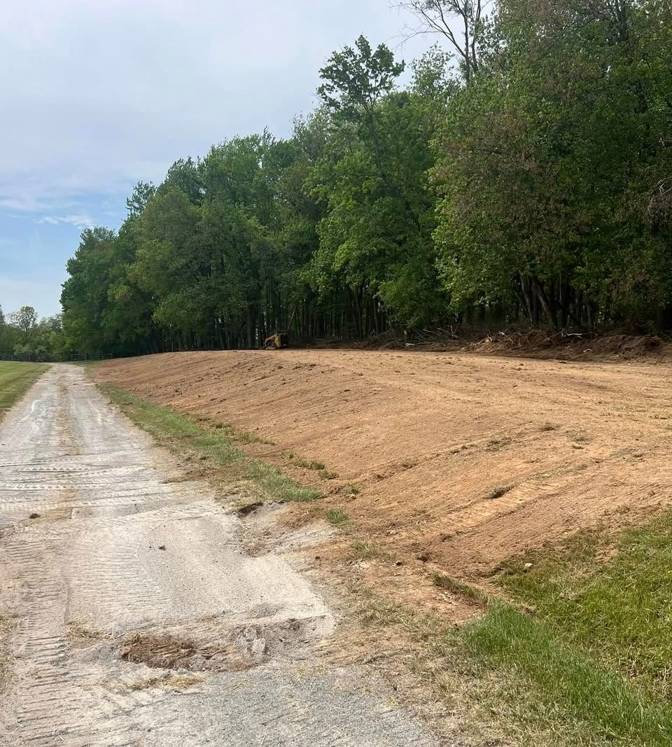 Dirt road next to cleared, graded land, bordering a tree line. Overcast day.