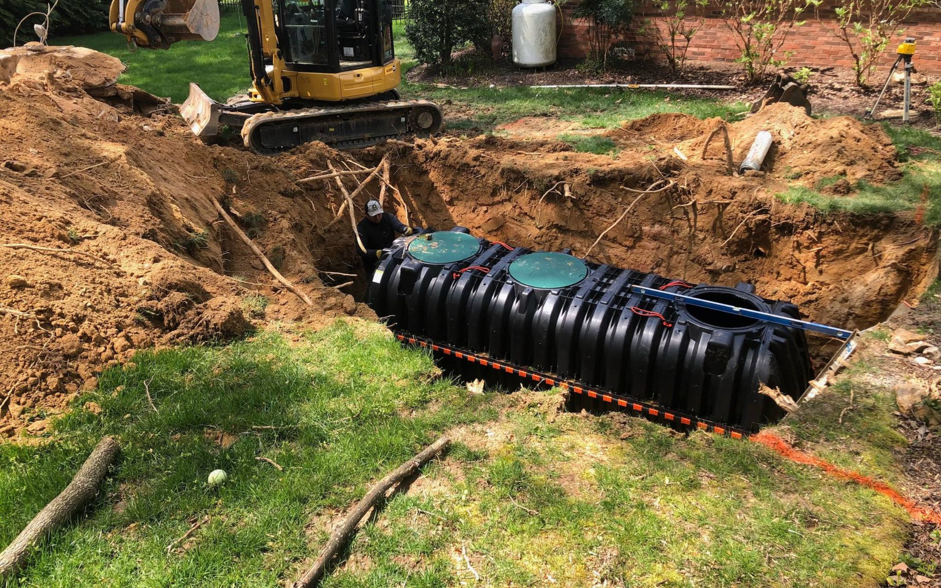 Excavator installing a black septic tank in a yard, surrounded by dirt and grass.