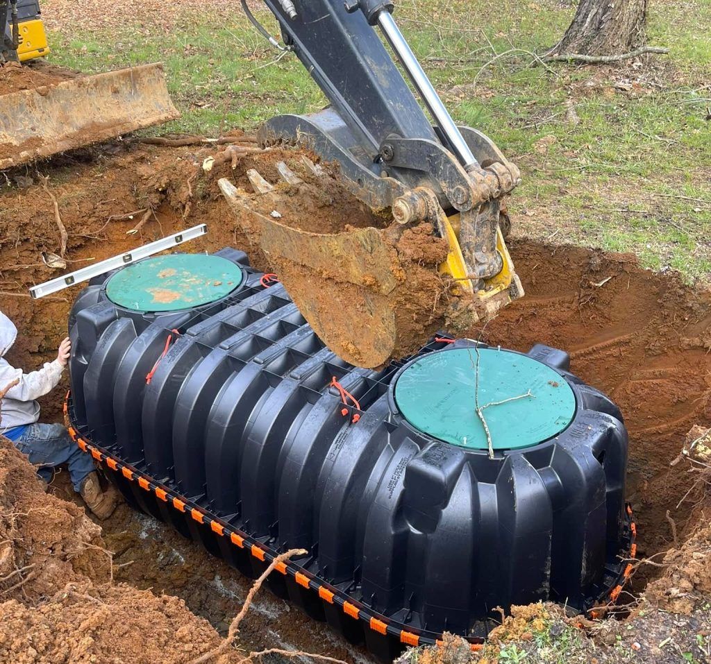 Excavator lowers black septic tank into a dug pit. Two green access lids are visible.