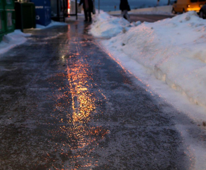 Icy sidewalk reflecting streetlights; snowbanks on the right. Pedestrians blurred in background.