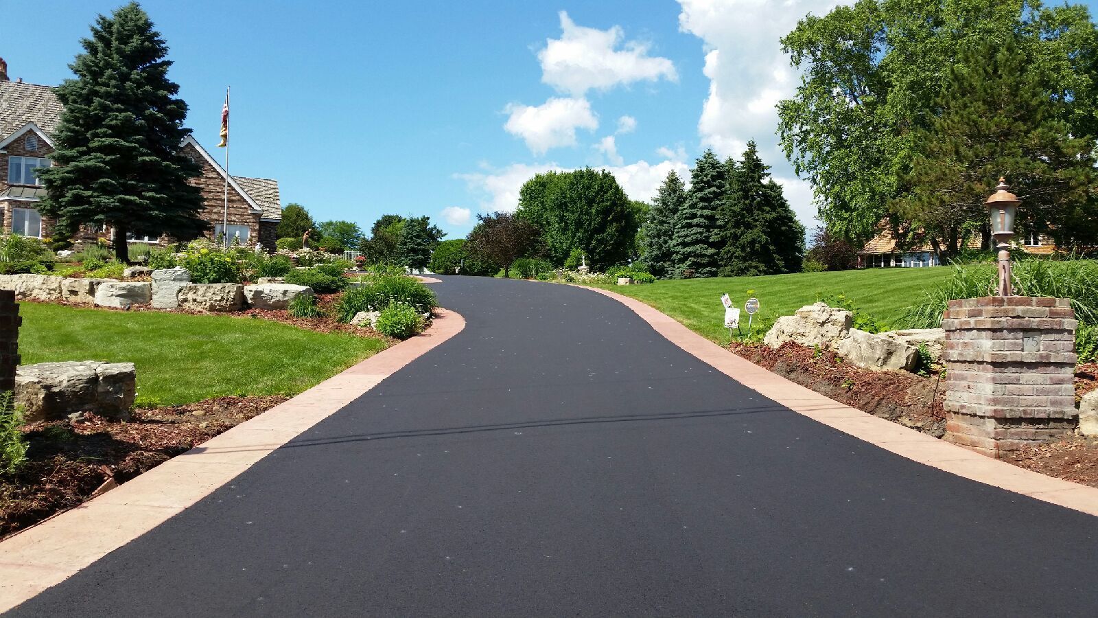 A paved driveway leads toward a stone house with a lush lawn, trees, and stone landscaping accents under a blue sky.