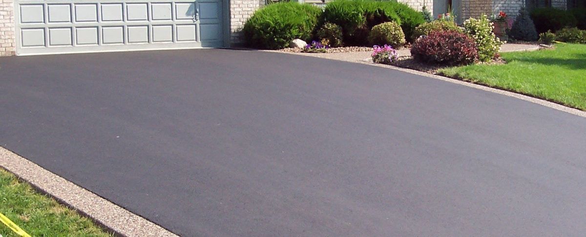 A black asphalt driveway in front of a house, with a garage door and landscaping.