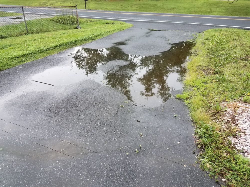 A large, stagnant puddle of water sits in the center of a weathered, cracked asphalt driveway near a road.