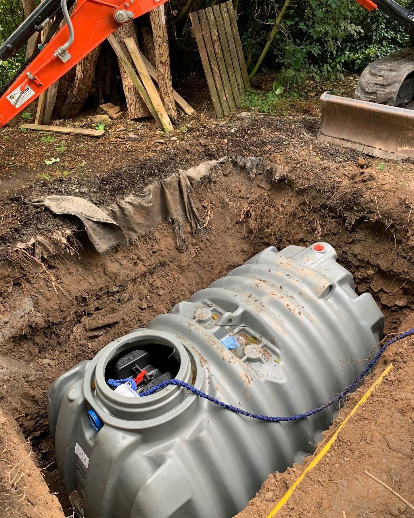 Gray septic tank in an excavated hole, excavator arm visible.