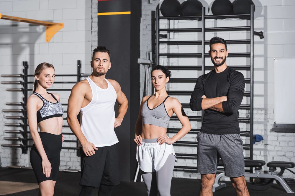 Cuatro personas vestidas con ropa deportiva están de pie una al lado de la otra en un gimnasio con espalderas al fondo.
