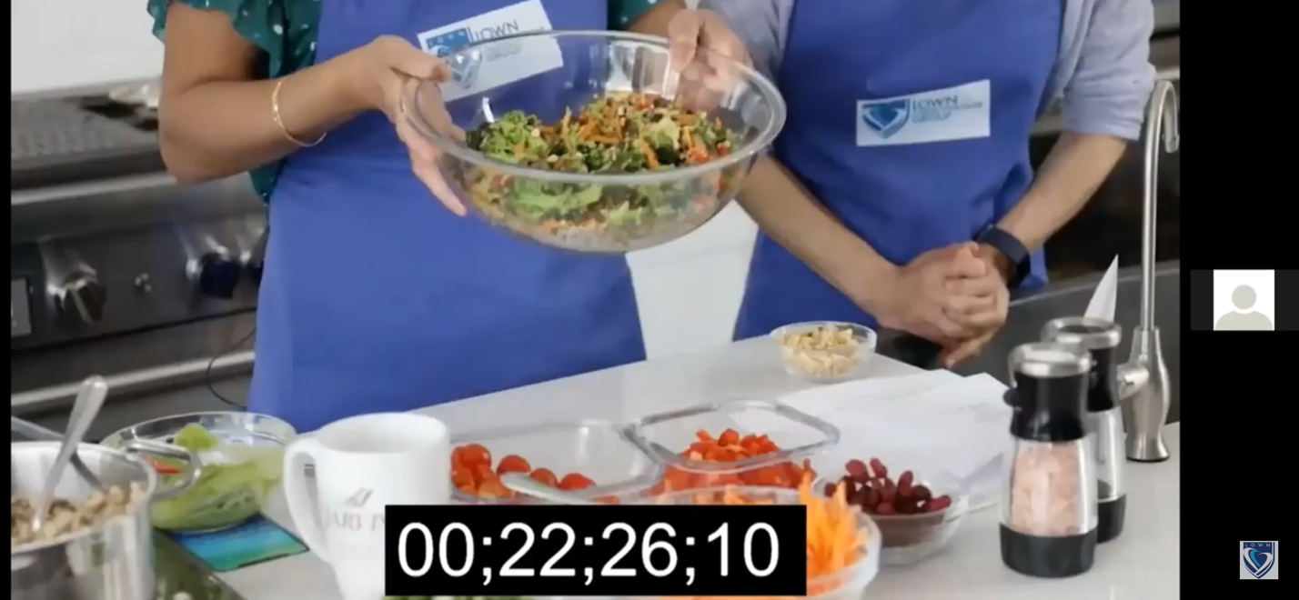 Two people in blue aprons hold a salad bowl, preparing food in a kitchen.
