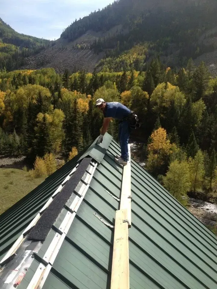 Roofer works on a green metal roof in a mountain setting with fall foliage.