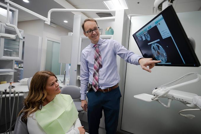 A man is talking to a woman in a dental chair while pointing at a screen.