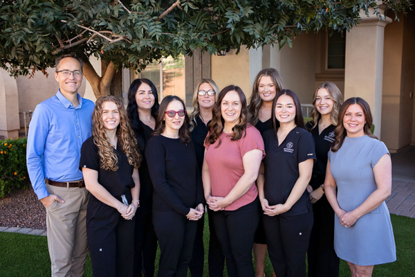 Group of people posing outdoors, smiling. Several women in scrubs, one man in a blue shirt.
