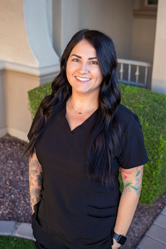 A woman in a black scrub top is smiling and standing in front of a building.