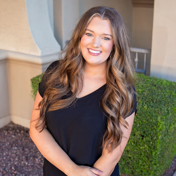 Woman in a black dress smiles outside, arms crossed, long wavy brown hair.