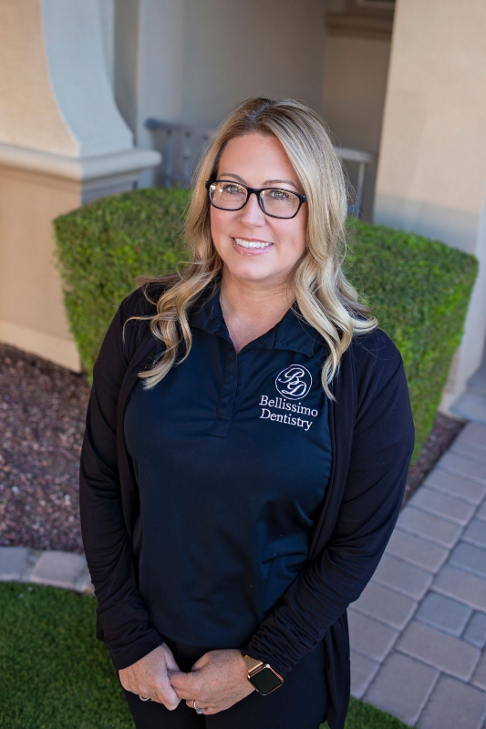 Woman wearing glasses and a black polo shirt, smiling, in front of a building with greenery.
