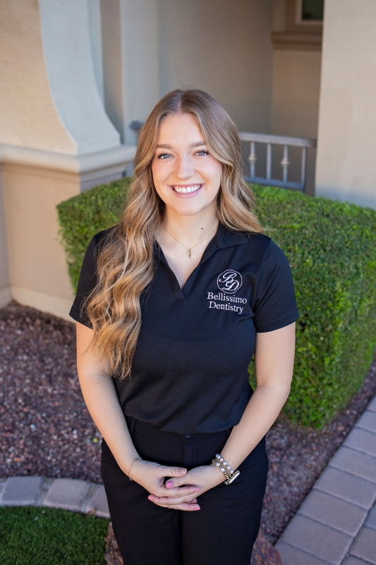 Woman with long wavy hair smiles, wearing black scrubs, standing outside a building.