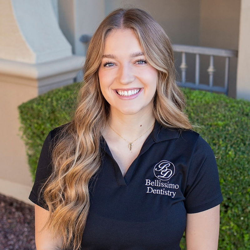 Woman with long wavy hair smiles, wearing black scrubs, standing outside a building.