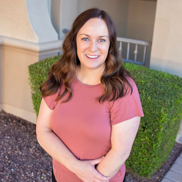 Woman in pink shirt smiles outdoors with arms crossed.