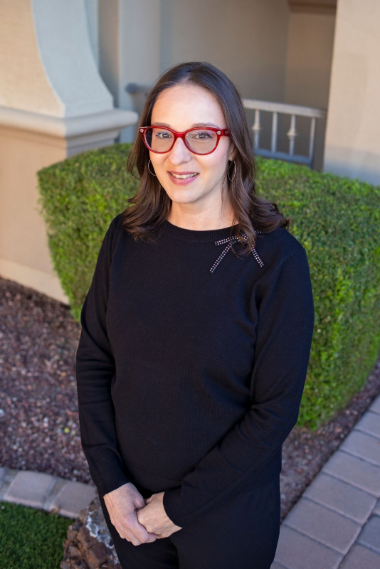 Woman with glasses in a black sweater standing by a trimmed hedge.