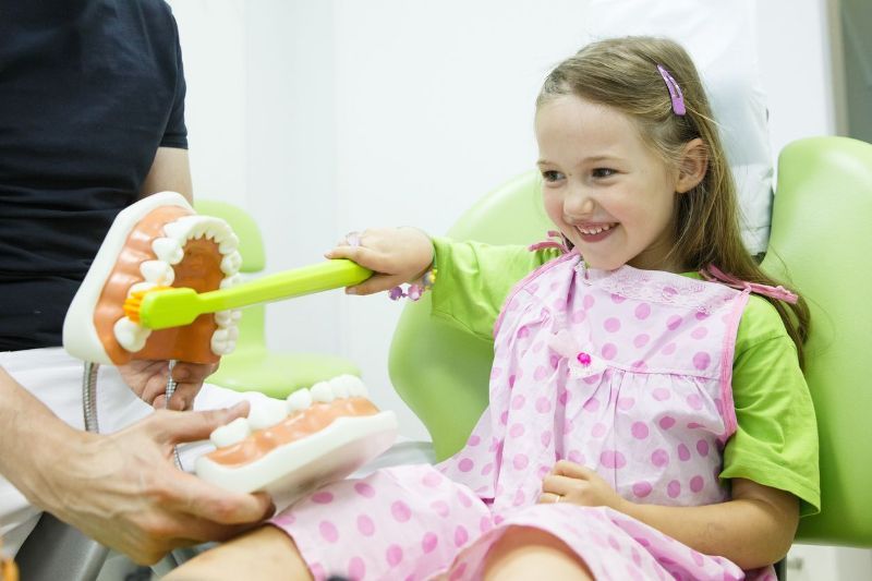 A little girl is sitting in a dental chair brushing her teeth with a toy toothbrush.