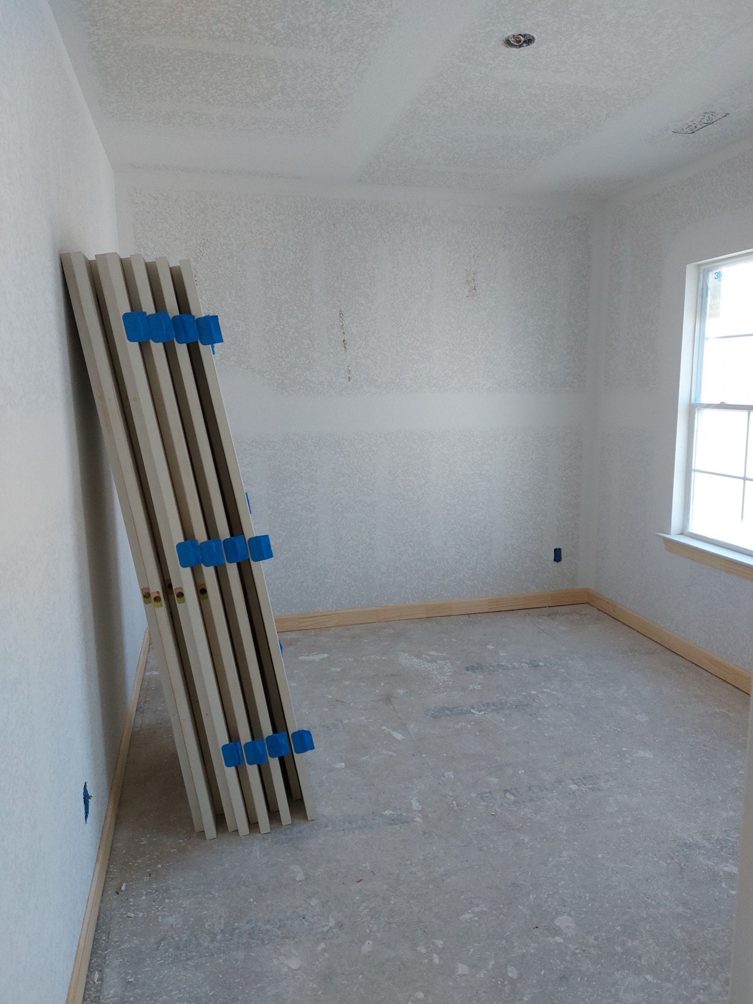 Empty room under renovation with stacked boards leaning against the wall and a window on the right.