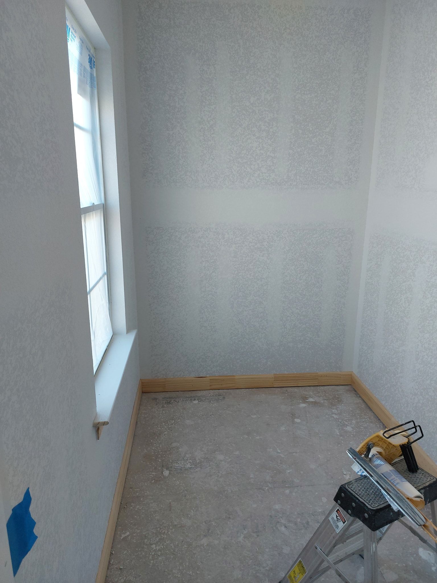 Small unfinished room with white walls, window, bare floor, and paint supplies in the corner.