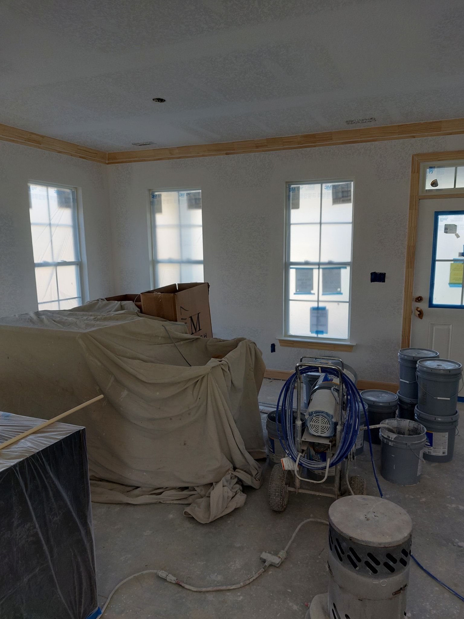Unfinished room with covered furniture, paint equipment, and tools by windows and a door.