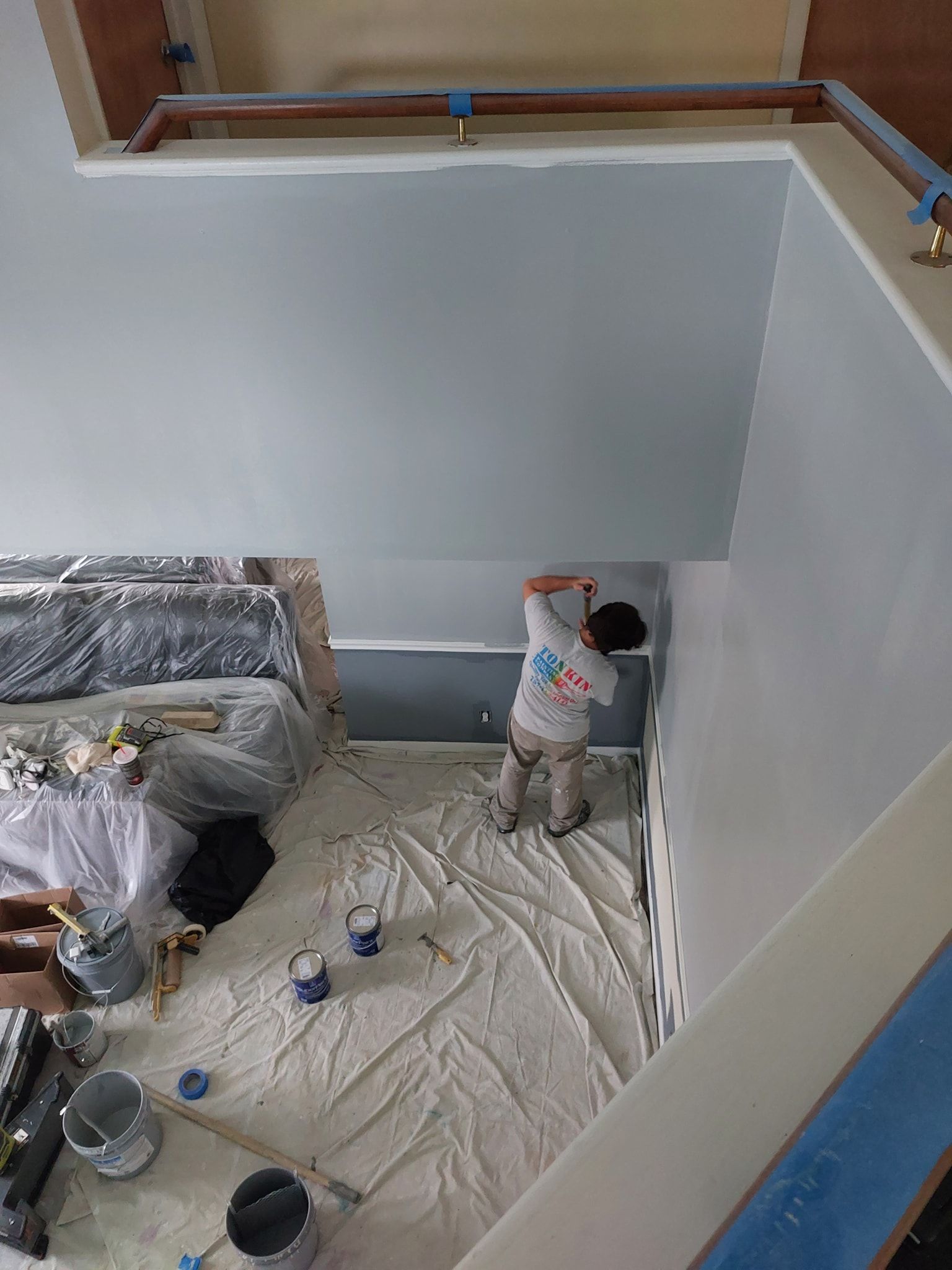 Worker painting a white interior wall in a room under renovation, with tools and drop cloths on the floor.