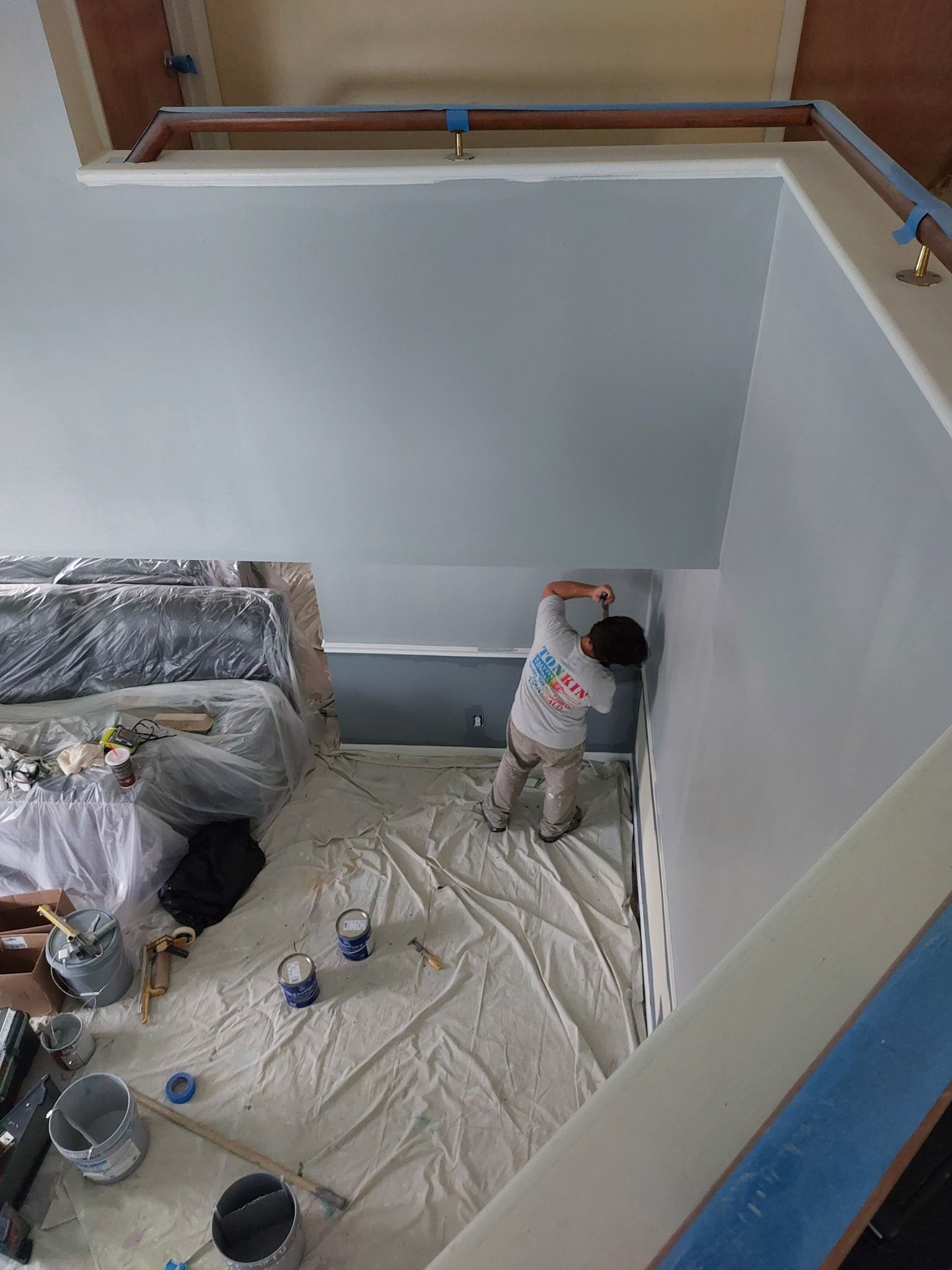Painter working on a room interior with white walls, tarps on the floor, and a blue railing above.