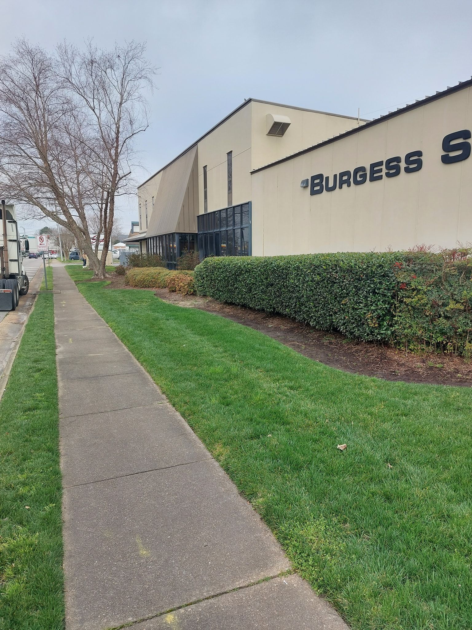 Sidewalk beside a beige industrial building with a hedge, grass, and leafless tree on an overcast day