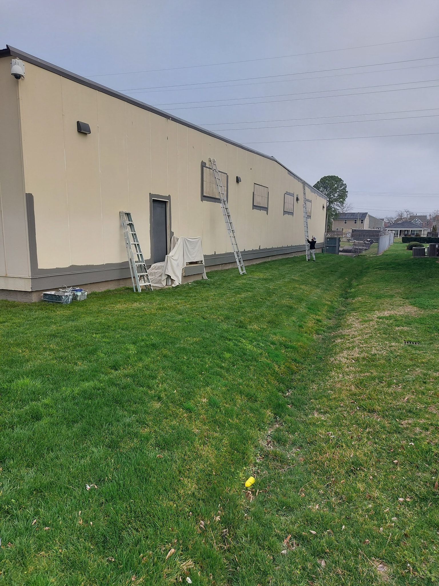 Long beige building beside a grassy field, with ladders leaning against the wall under an overcast sky.