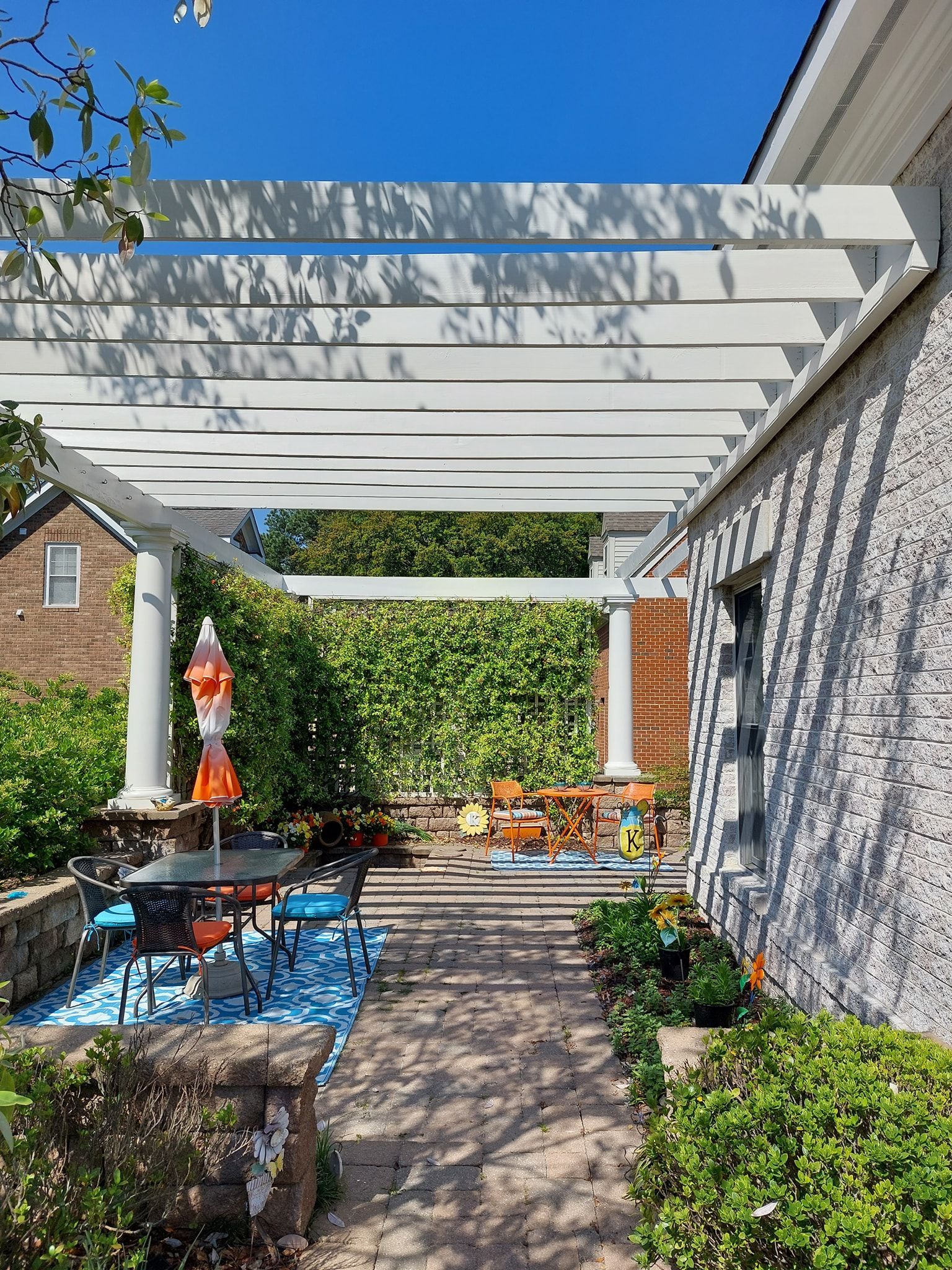 Patio with white pergola, brick wall, blue rug, potted plants, and outdoor seating in bright sunlight