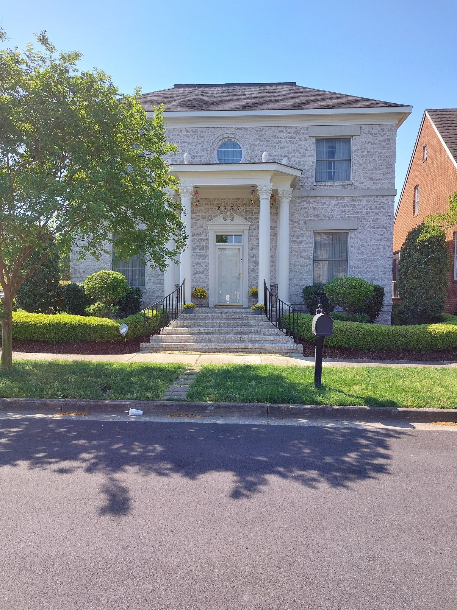 Large white two-story house with front steps, columns, and a manicured lawn on a sunny street.