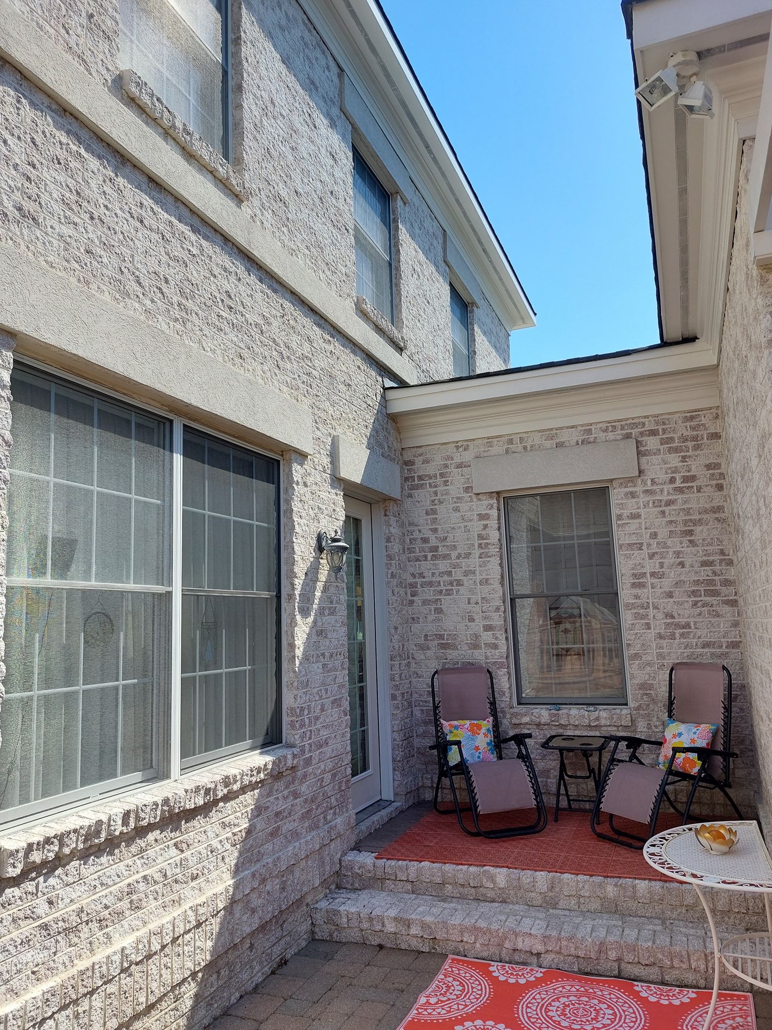 Small stone patio between houses with two chairs, a table, potted plants, and a screened window.