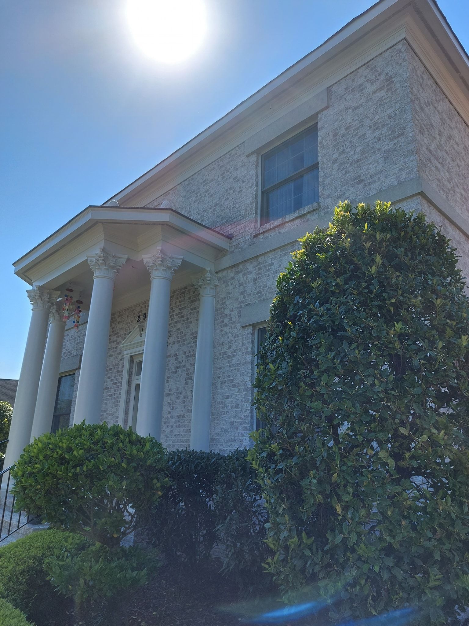 Grand white-columned house with stone facade, bright sun, and trimmed bushes in the foreground