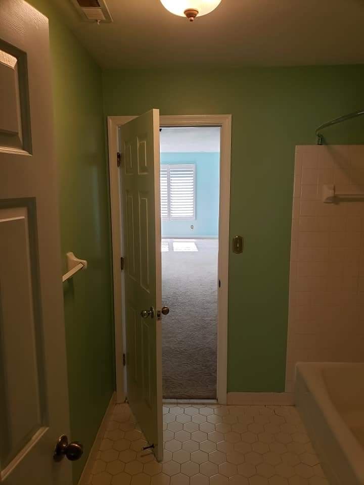 Green bathroom with tiled floor, open door to a carpeted bedroom, and a white shower curtain on the right.
