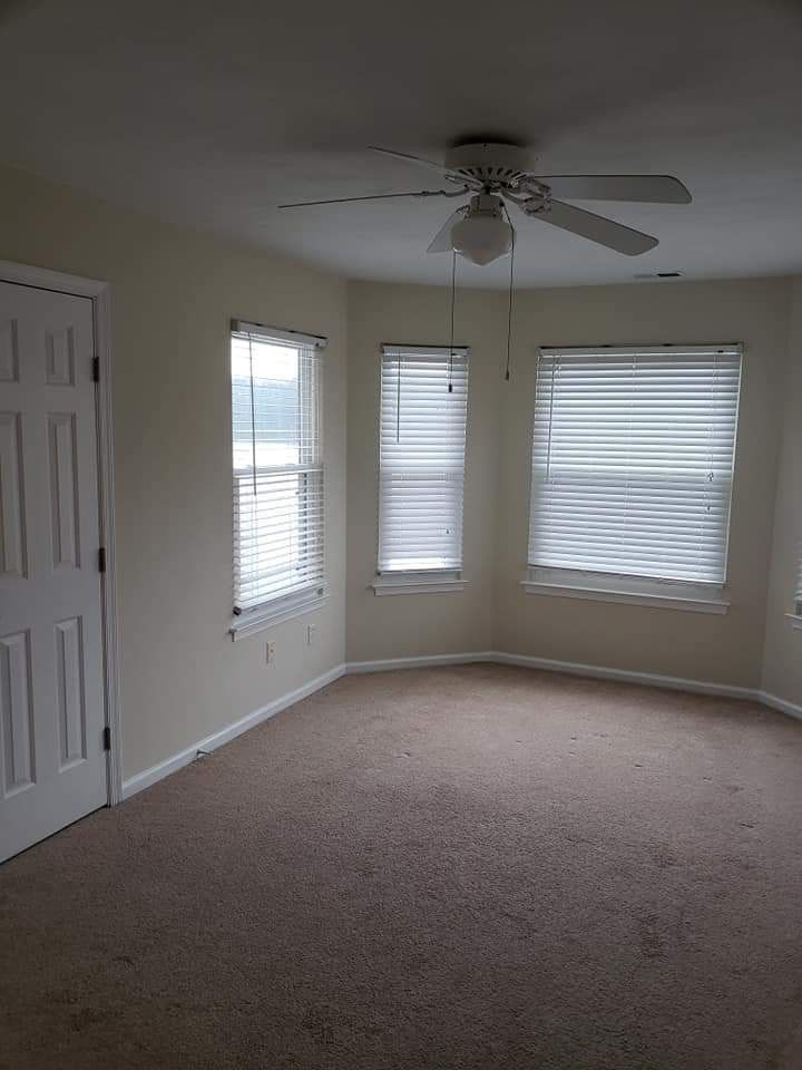 Empty beige bedroom with carpet, three windows, and a ceiling fan; closet doors on the left.