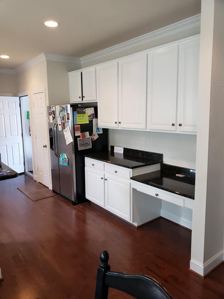 White kitchen with black countertops, refrigerator, and built-in desk area along a hardwood hallway