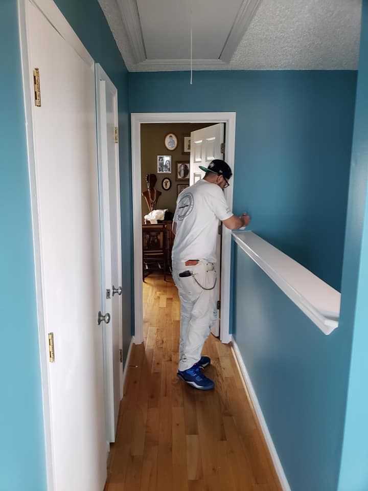 Person in white work clothes painting a blue hallway railing inside a home.
