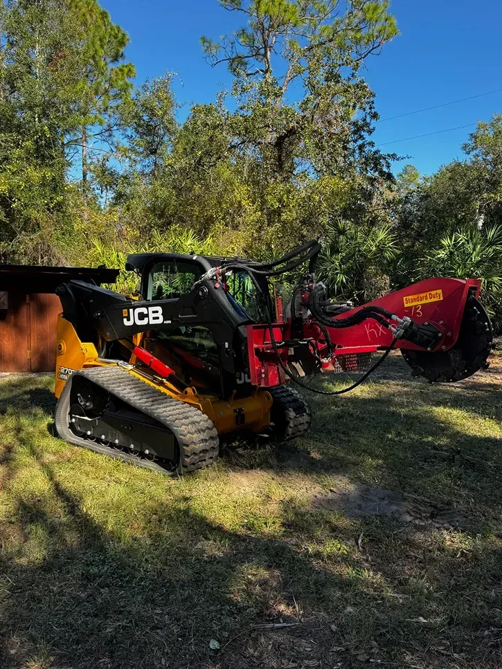 Black and yellow JCB skid steer with a red stump grinder, in a grassy yard.