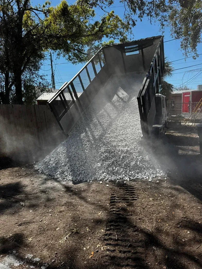 A dump truck unloading gravel on a dirt road, creating a cloud of dust.