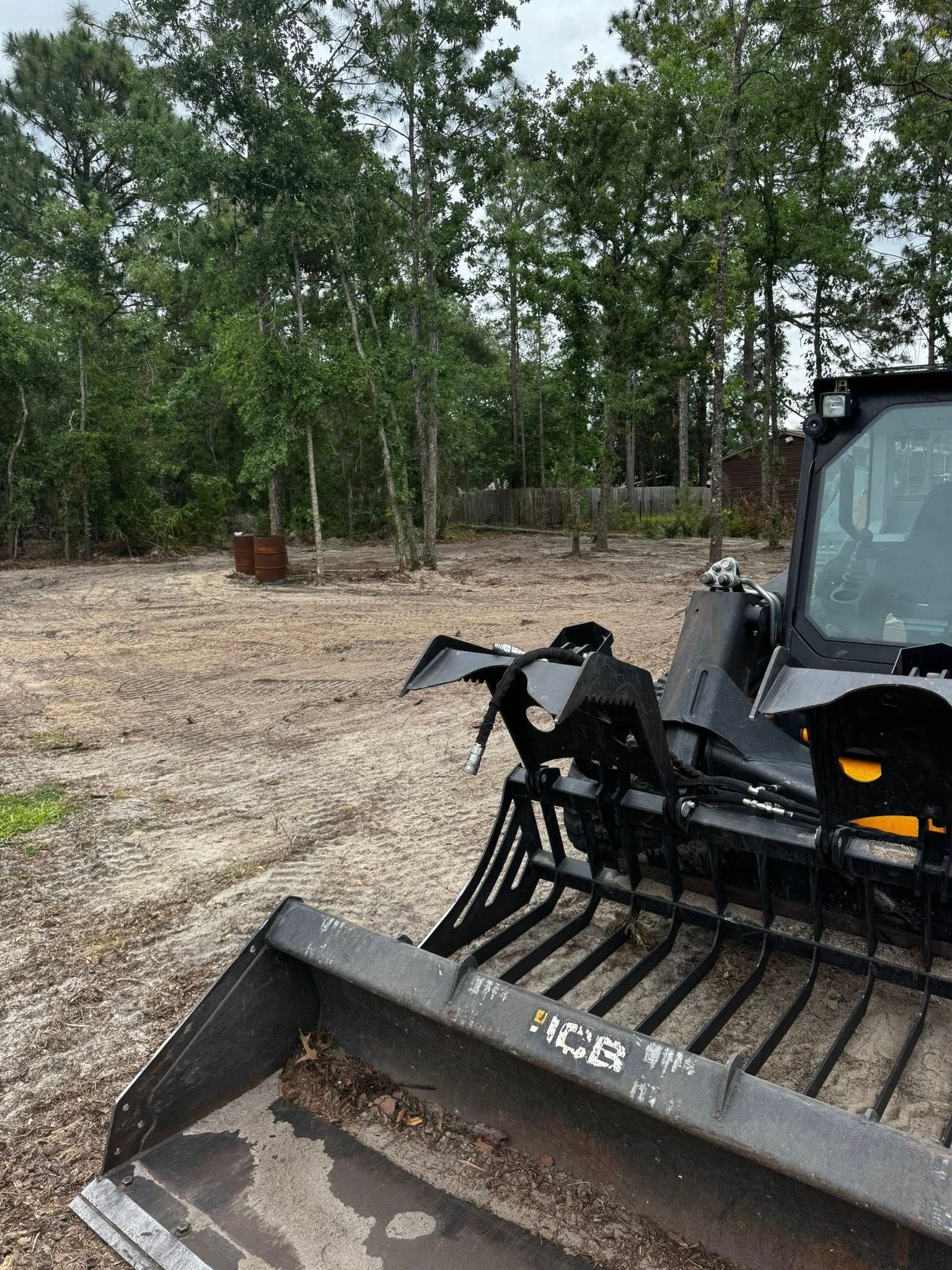 A skid steer with a grapple attachment on a dirt lot with trees in the background.