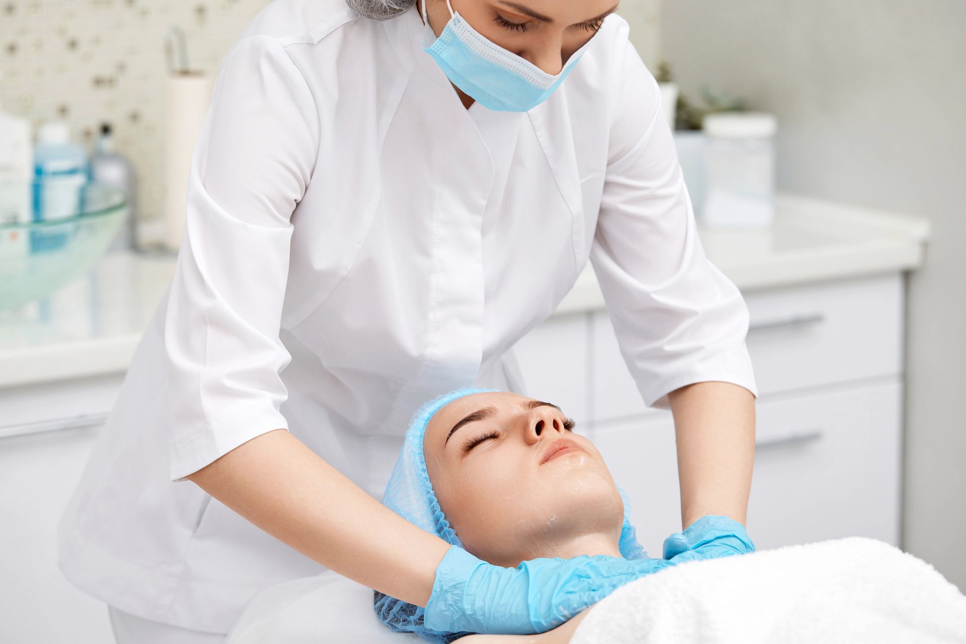 Un profesional con uniforme blanco y guantes azules realiza un masaje facial a una persona que está tumbada en una clínica.