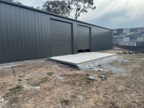 A large black metal building with two roller doors is sitting in the middle of a dirt field.